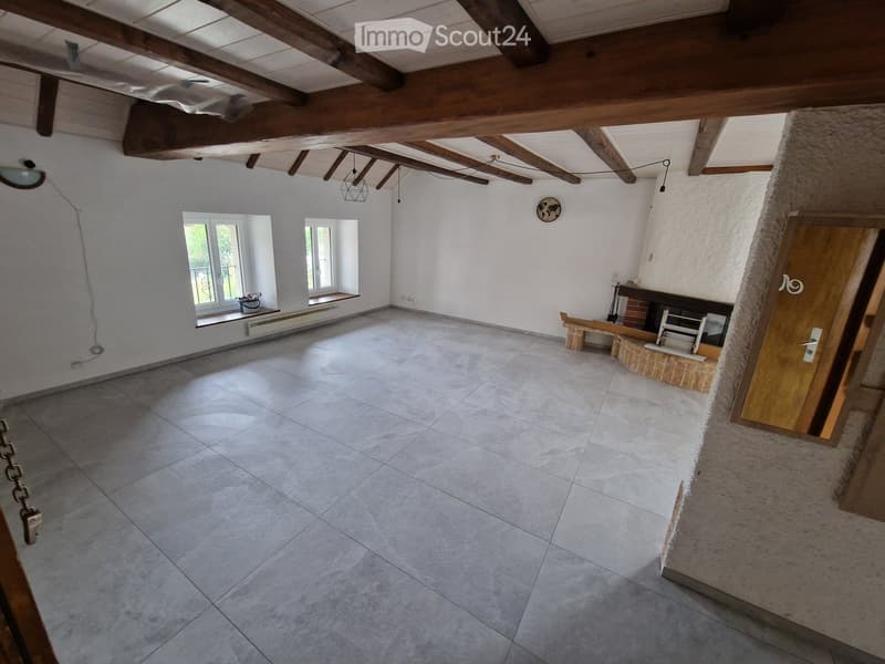 Empty room with exposed wooden beams, tiled floor, and white walls. Features a fireplace, windows with shutters, and a small bench.