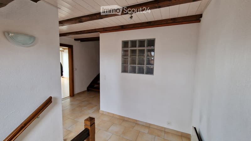 A hallway with white walls, tiled floor, and wooden ceiling beams. A staircase leads upwards, and a glass block window is visible on one wall.