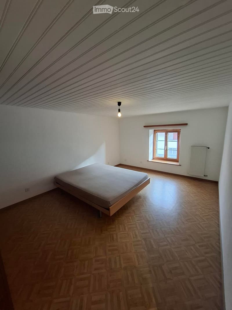 Bedroom with wooden floor, slanted ceiling, single bed, window with wooden frame, radiator, and pendant light.