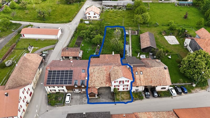 Aerial view of a residential building with multiple sections, red-tiled roofs, solar panels, and a parking area with several cars.