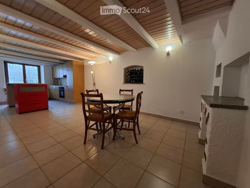 Open-plan dining and kitchen area with tiled flooring, wooden ceiling beams, and modern kitchen cabinets. Includes a round dining table with four chairs, a wine rack, and a floor lamp.