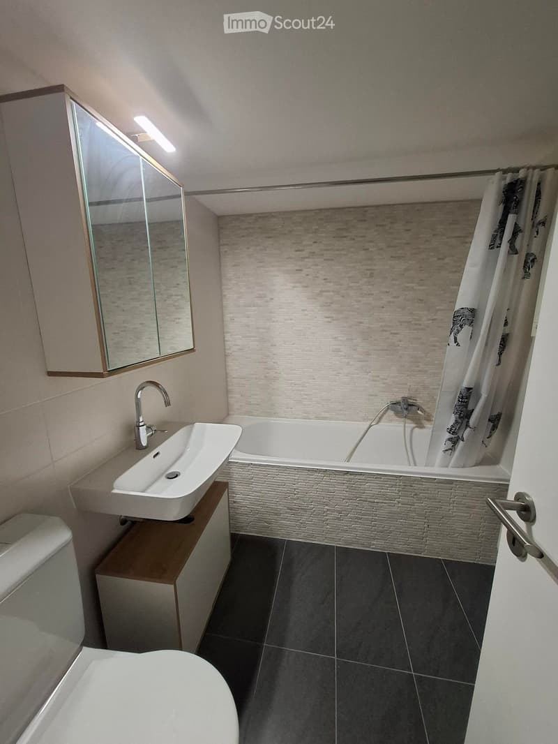 Modern bathroom with a white sink, bathtub, toilet, and a large mirror with wooden frame. The walls are tiled in a light beige color, and the floor is dark grey tiles.