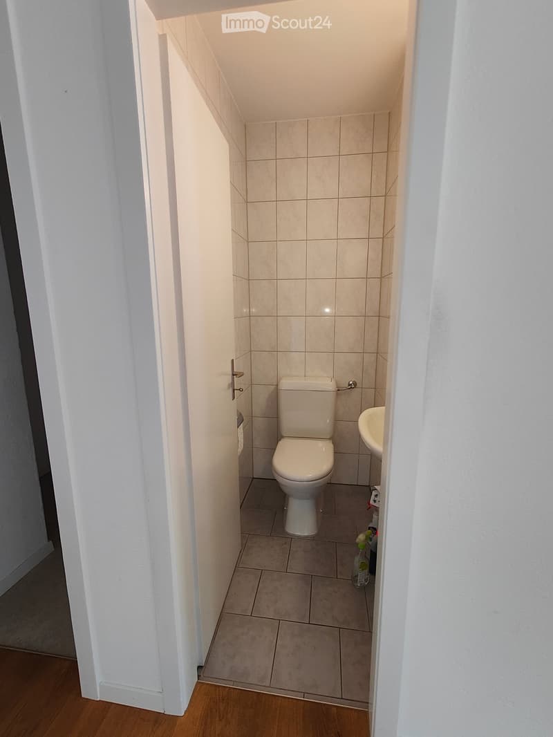 Small bathroom with white tiled walls and floor, toilet, sink, and cleaning supplies visible.