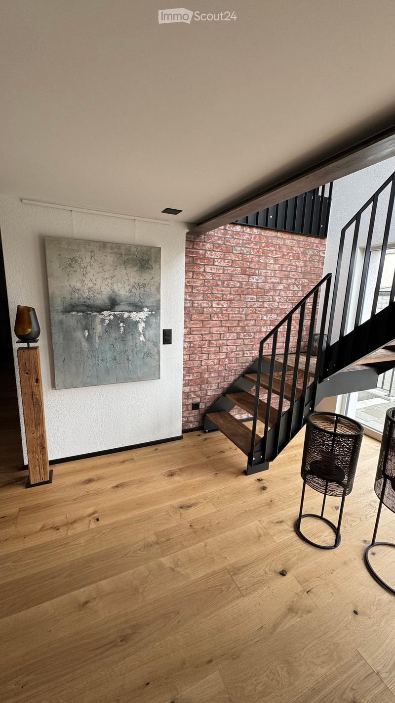 Modern hallway with wooden flooring, a staircase with metal railing, and a brick accent wall. Features include a large abstract painting and a decorative wooden stand with a vase.
