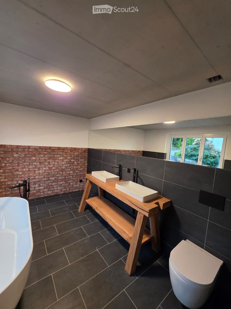 Modern bathroom with dark tiled floor, brick accent wall, white bathtub, toilet, and a wooden shelf holding two sinks.