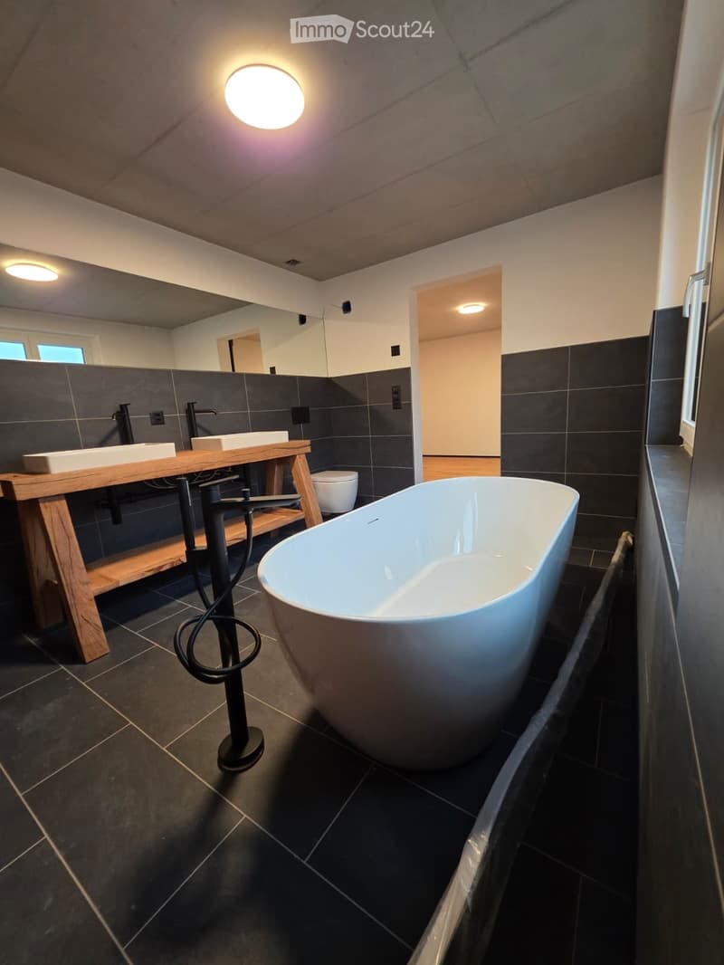 Modern bathroom with dark tiled walls and floor, a freestanding bathtub, a wooden vanity with two sinks, and a toilet. The space features sleek, contemporary fixtures and ample lighting.