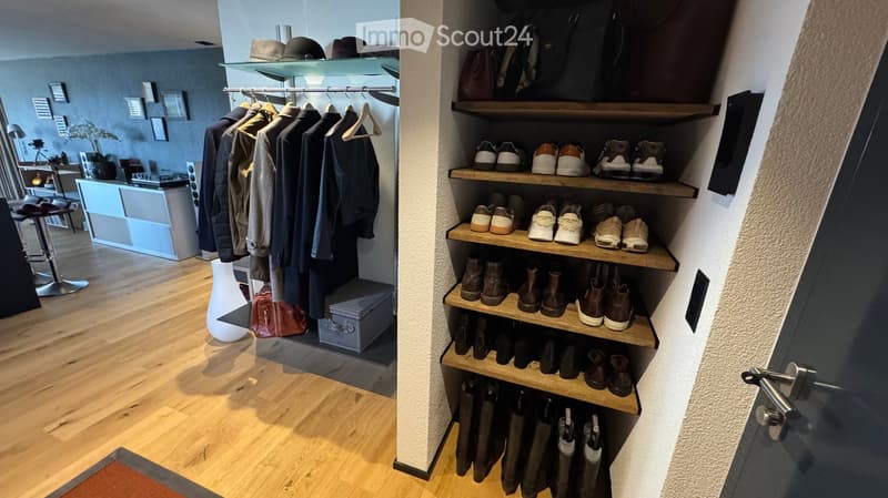 A well-organized entryway with a wooden floor, a built-in wardrobe with hanging clothes, a shoe rack with various footwear, a small table with a vase, and picture frames on the wall.