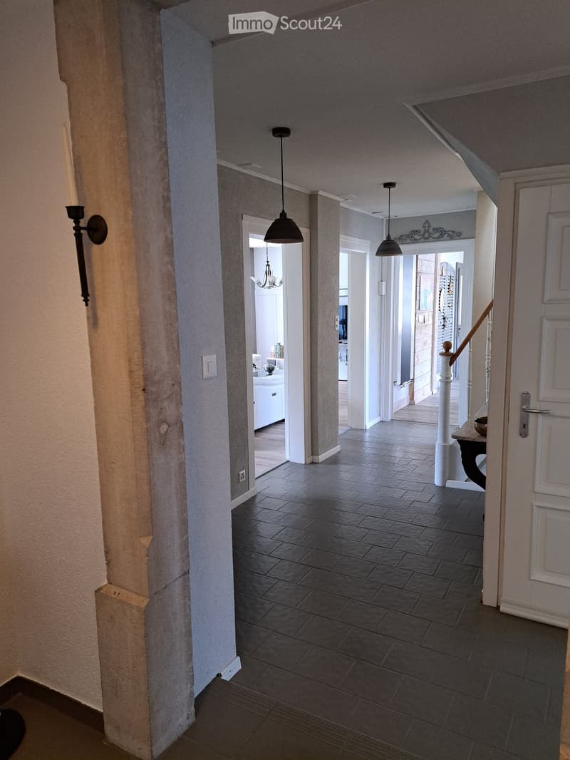 A spacious hallway with dark tiled flooring, white walls, and several hanging pendant lights. The hallway features an open staircase leading upwards, with visible rooms on either side, including a bathroom and a living area.