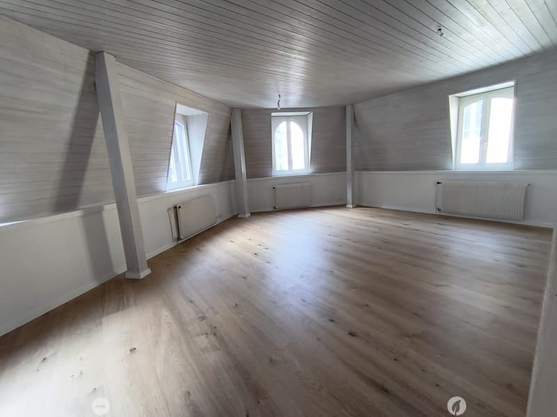 Empty room with wooden flooring, white paneled walls, and multiple windows allowing natural light. Radiators are visible along the walls.