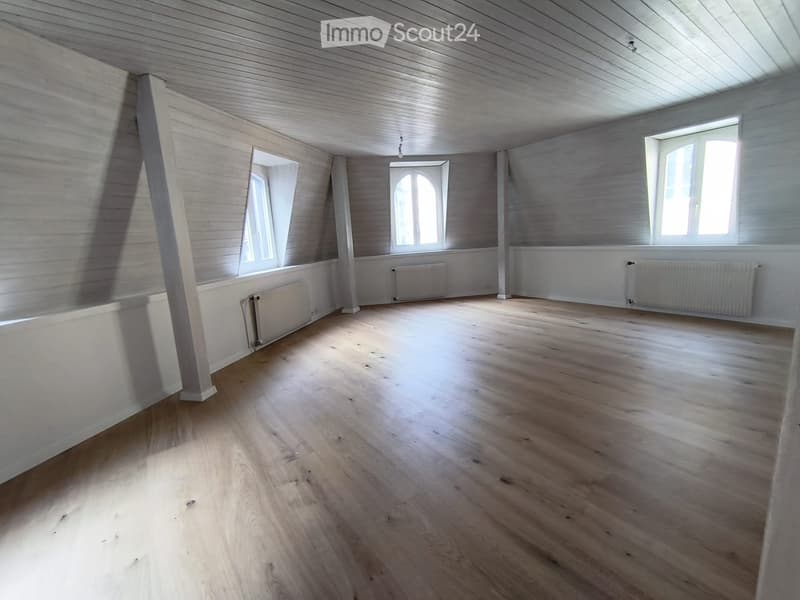 Empty room with wooden flooring, white paneled walls, and multiple windows allowing natural light. Radiators are visible along the walls.