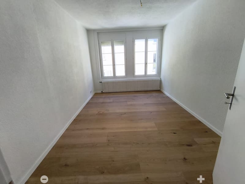 Empty room with light wooden flooring, white walls, and a large window with white frames and blinds. A radiator is visible below the window.