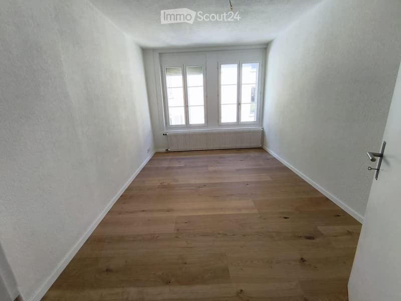 Empty room with light wooden flooring, white walls, and a large window with white frames and blinds. A radiator is visible below the window.