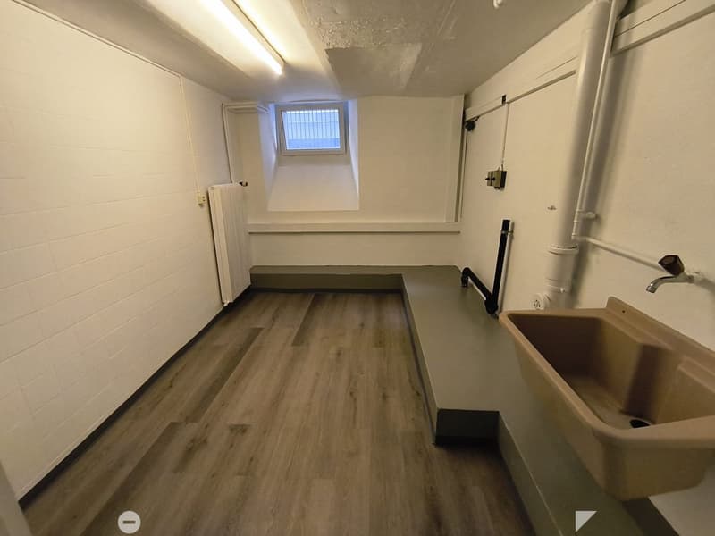 Empty room with wooden flooring, white brick walls, a small window with bars, a radiator, and a sink with a faucet.