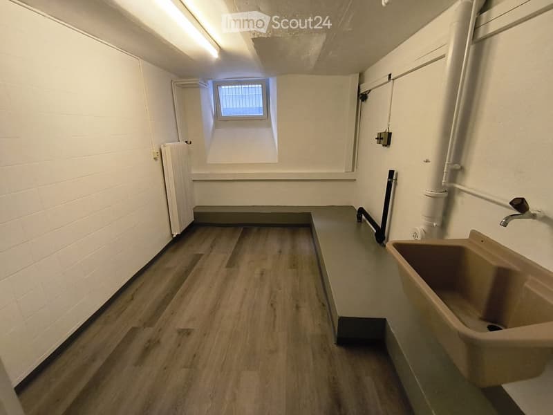 Empty room with wooden flooring, white brick walls, a small window with bars, a radiator, and a sink with a faucet.