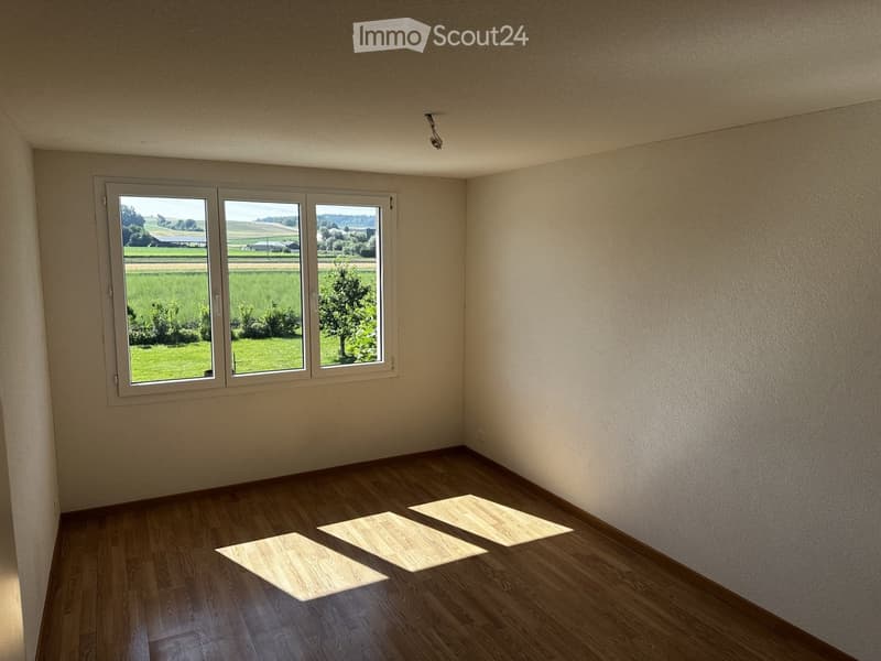 Empty room with wooden flooring and large windows overlooking a green field. No furniture or equipment present.