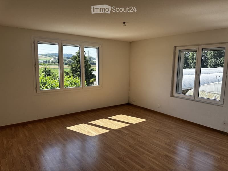Empty room with wooden flooring and large windows offering a view of greenery and a greenhouse. No furniture or equipment present.