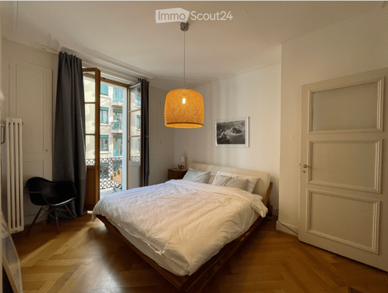 Cozy bedroom with a large bed, white bedding, a black chair, a nightstand, and a pendant light hanging from the ceiling. The room features wooden flooring and large windows with curtains, offering a view of the street.