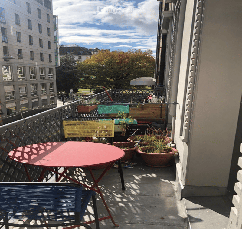 Balcony with a red round table, blue chair, and several potted plants arranged on the railing and floor.