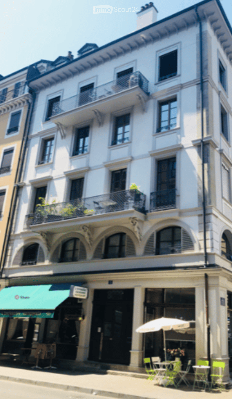 Multi-story building with white facade, multiple balconies, and a small outdoor seating area with tables and chairs.