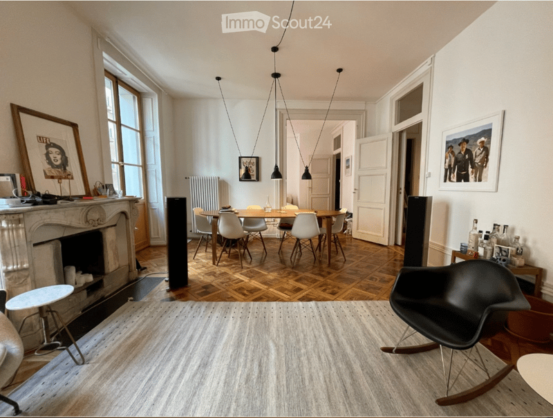 A modern, well-lit living space with a fireplace, a dining area, and a comfortable seating area. The room features a mix of contemporary and classic furniture, including a rocking chair, a coffee table, and a dining table with chairs.