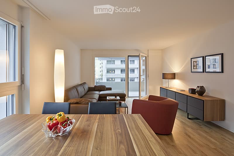 Modern living room with a sectional sofa, coffee table, dining table, chairs, and a balcony view. The room features wooden flooring, large windows, and contemporary decor including a lamp and a cabinet.