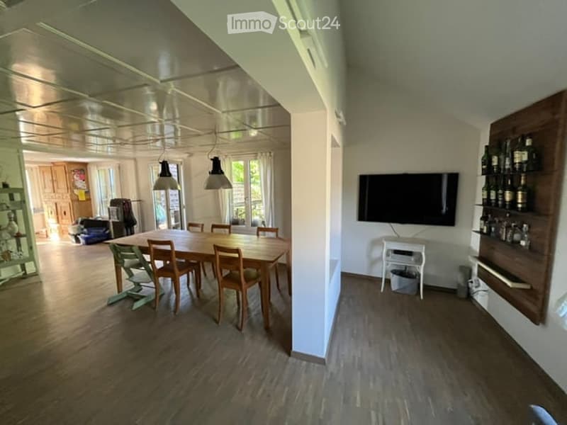 Bright dining area with a wooden dining table and chairs, a flat-screen TV mounted on the wall, and a wooden shelf with bottles. The space features wooden flooring and pendant lighting.