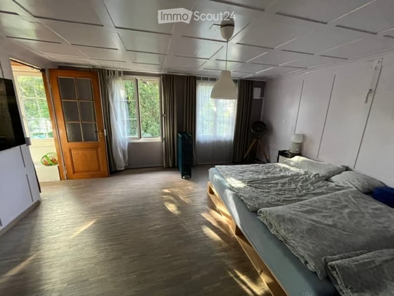 Bright bedroom with wooden flooring, large windows with curtains, a double bed with grey bedding, a nightstand with a lamp, and a small table with a fan nearby.