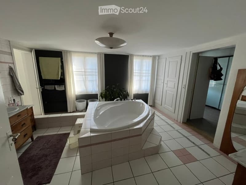 Bathroom with a large bathtub, sink, mirror, and a plant near the window. The room features tiled flooring and walls, with a curtain covering the window.
