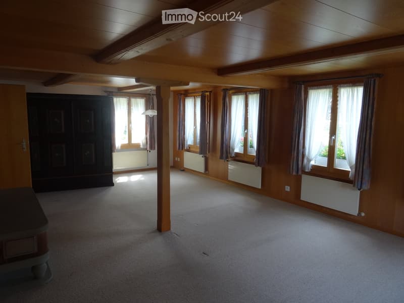 Empty room with wooden ceiling beams, large windows with curtains, carpeted floor, and a central support column.