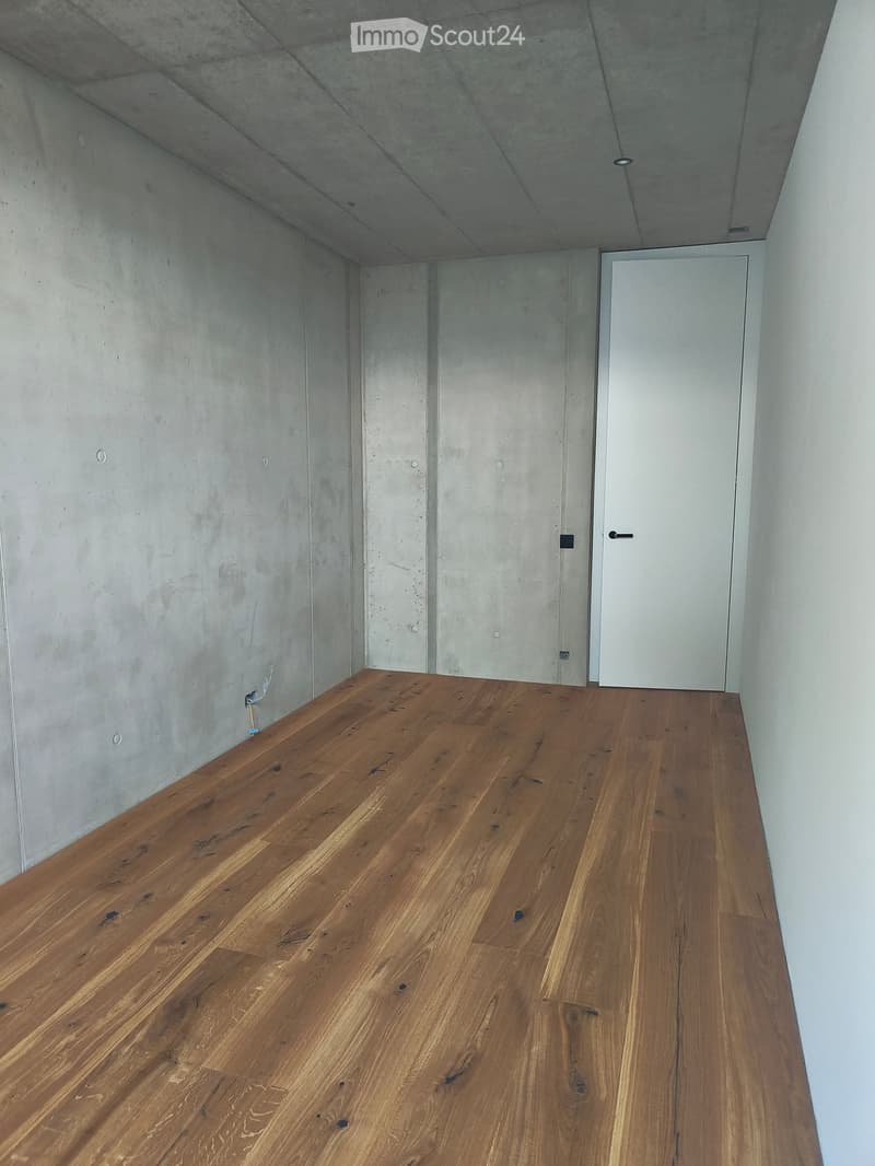 Empty room with polished wooden flooring and concrete walls, featuring a single white door.