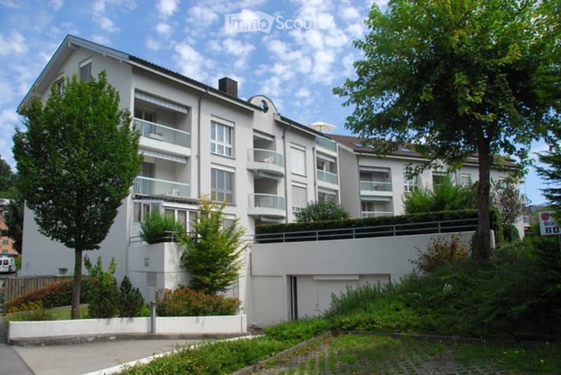 Modern multi-story apartment building with white facade, multiple balconies, and surrounding greenery.