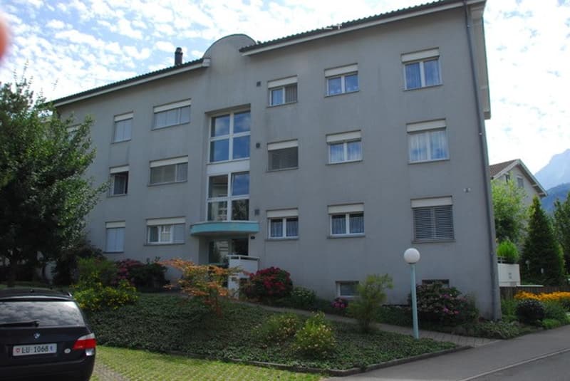 Multi-story residential building with a modern facade, large windows, and a landscaped front yard with flowers and shrubs.