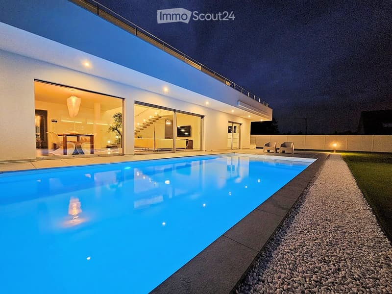 Modern two-story house with a large swimming pool in the foreground, illuminated at night. The house features large glass doors leading to a well-lit interior, with visible furniture and decor inside.
