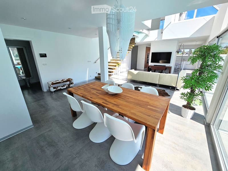 Modern dining area with a wooden dining table, white chairs, a white sofa, a television, and a potted plant. The space features large windows and a contemporary chandelier.