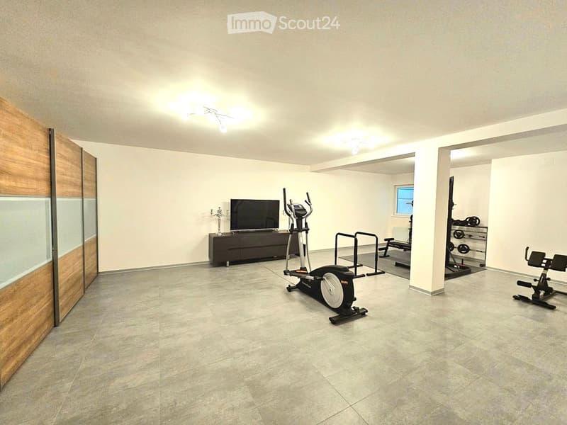 A spacious, well-lit room with tiled flooring and modern lighting. It features a television on a cabinet, an exercise bike, a bench, and various weights, creating a multifunctional space for relaxation and fitness.