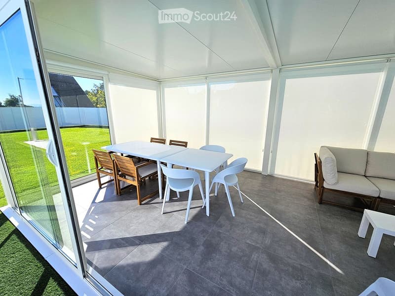 Modern enclosed patio with large glass walls, grey tiled floor, white sofa, dining table, and chairs.