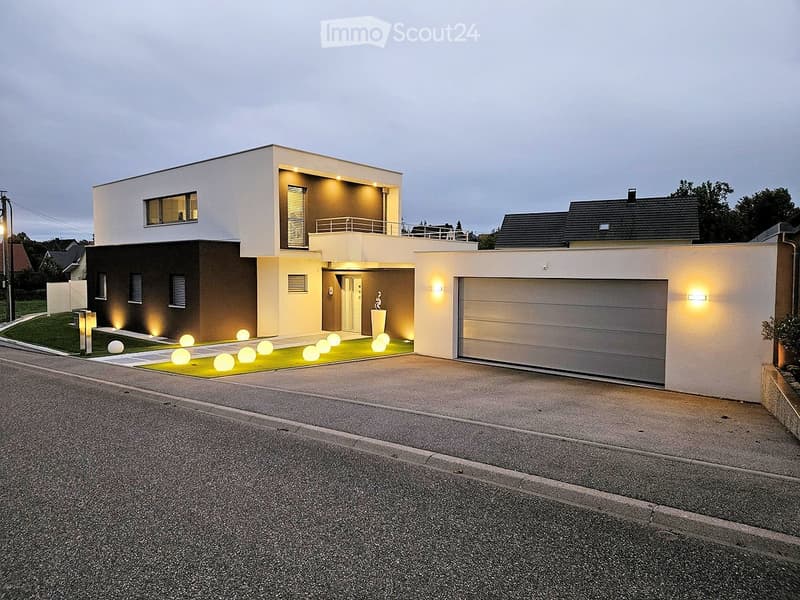 Modern two-story house with a combination of white and dark brown facades, illuminated entrance, garage, and small garden area with decorative lighting.