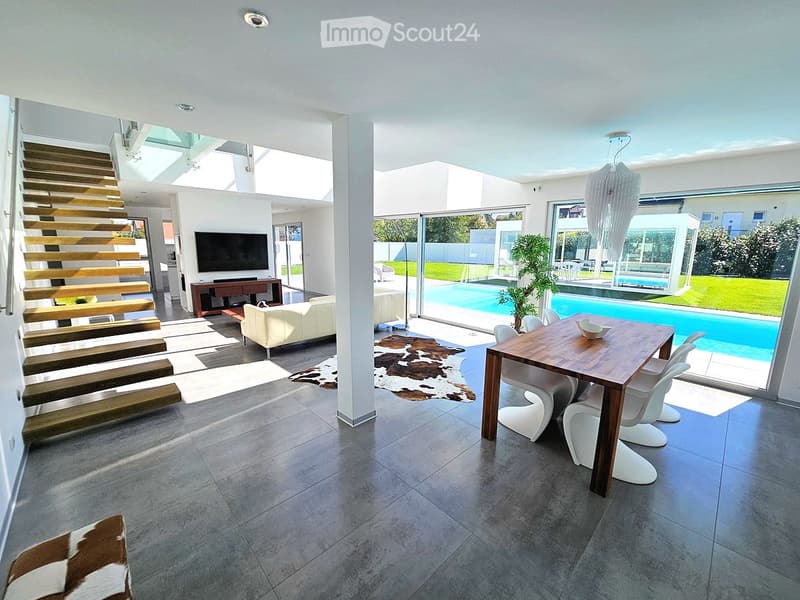Modern living and dining area with large windows, a swimming pool view, a white sofa, a wooden dining table with four white chairs, a television, and a staircase leading to an upper level.