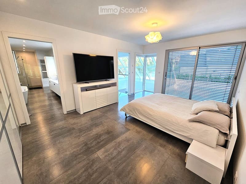 Modern bedroom with a large bed, flat-screen TV on a white stand, and large windows with blinds. The room features a tiled floor and a visible bathroom area through an open doorway.