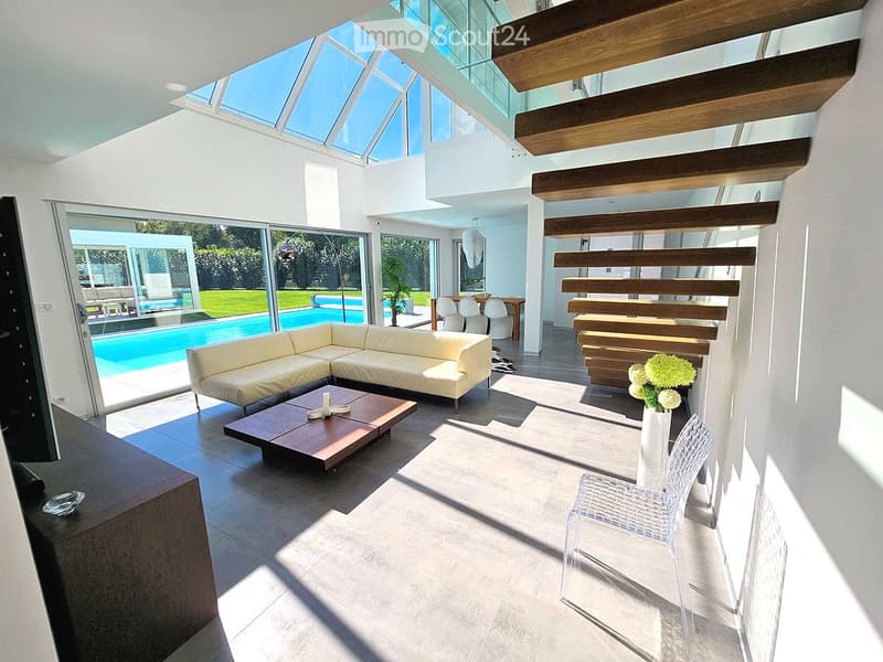Modern living room with large glass windows, a cream-colored sectional sofa, a wooden coffee table, a white vase with flowers, and a clear plastic chair. The room has a high ceiling with wooden beams and a skylight, leading to a pool area outside.