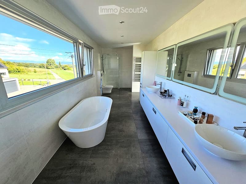 Modern bathroom with a large window offering a scenic view, featuring a freestanding bathtub, double sink vanity with mirrors, and a walk-in shower.