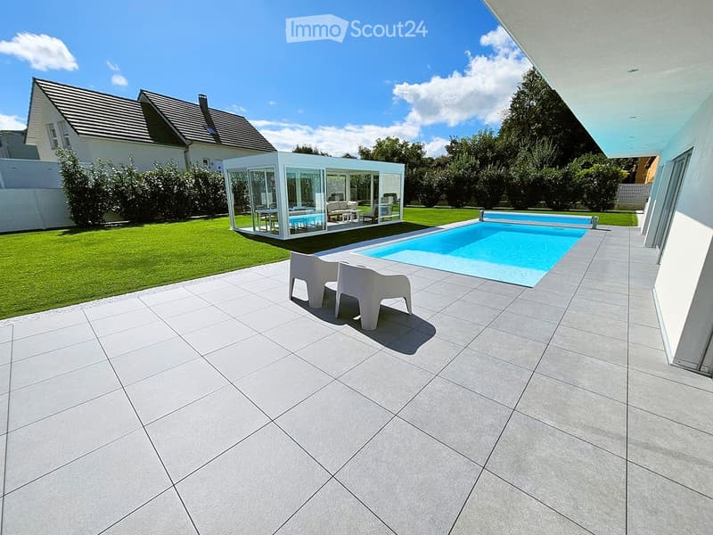 Modern outdoor area with a swimming pool, tiled patio, white lounge chairs, and a glass-walled pavilion with indoor seating.
