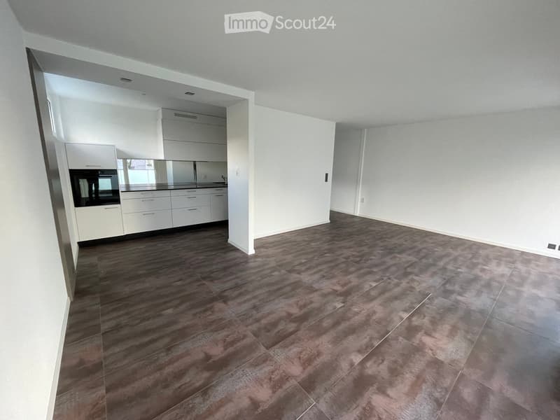 Empty room with light-colored walls, dark wood flooring, and an adjacent kitchen area with white cabinets and a built-in oven.