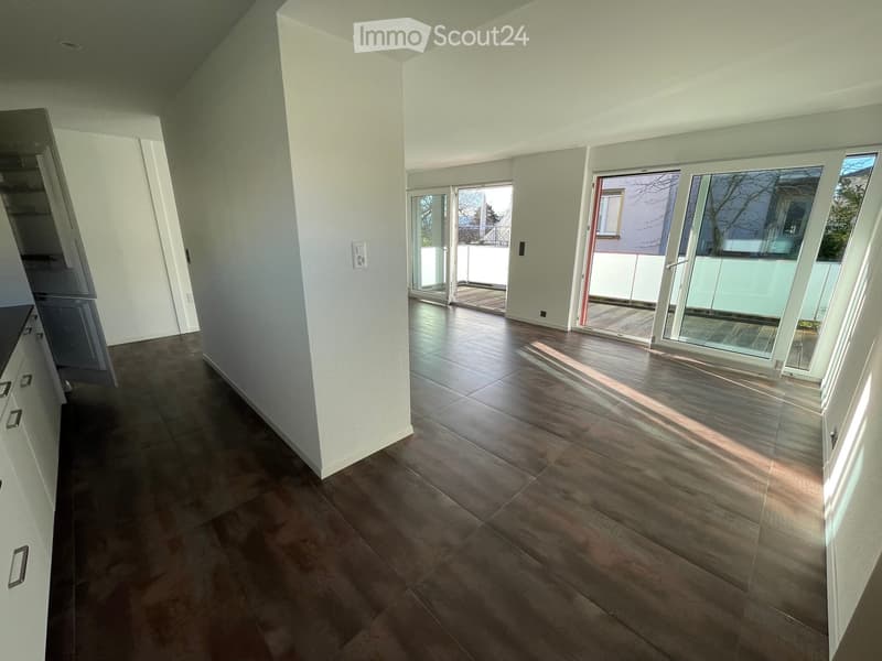 Bright, modern hallway with wooden flooring, white walls, and large glass doors leading to a balcony area.