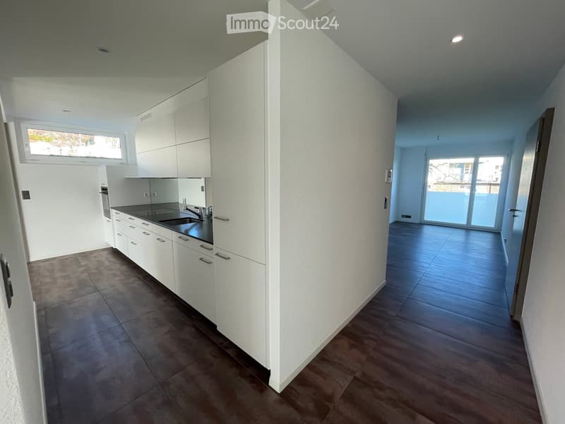 Modern, unfurnished kitchen and living area with white cabinets, dark countertops, and large windows. The space features tiled flooring in the kitchen and wooden flooring in the living area, with a sliding glass door leading to a balcony.