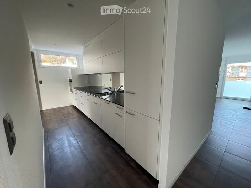 Modern kitchen with white cabinets, dark countertops, and a stainless steel sink. The space is well-lit with natural light from a window and has a tiled floor.