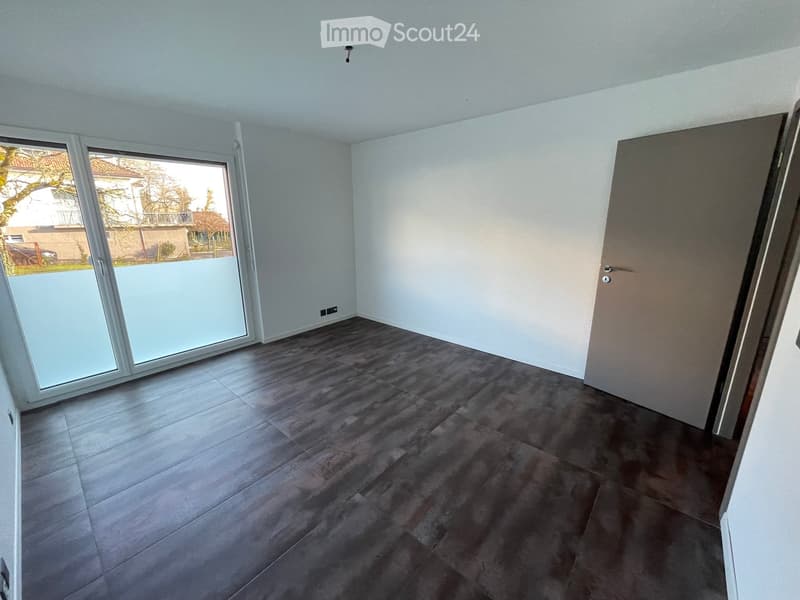 Empty room with dark grey tiled floor, white walls, large sliding glass door leading outside, and a closed door on the right side.