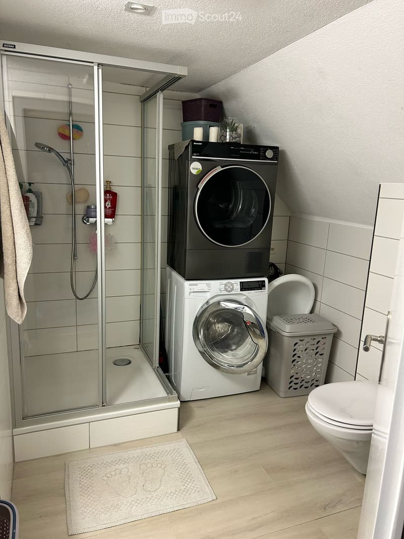 Modern bathroom with a glass-enclosed shower, a washing machine, a toilet, a laundry basket, and a towel on the floor.