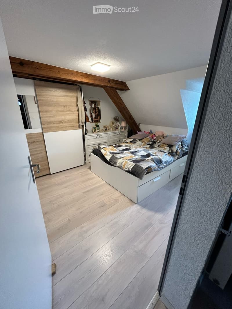 Cozy attic bedroom with wooden floors, slanted wooden ceiling beams, a white bed with a patterned quilt, a bedside table with a lamp, a mirror, and a wardrobe with a curtain.