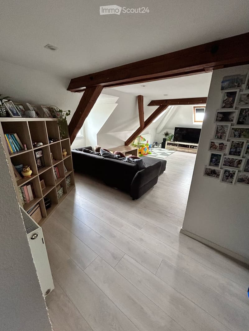Bright living room with wooden beams, white walls, and light-colored flooring. Features a dark-colored sofa, coffee table, bookshelf, television, and several picture frames on the wall.
